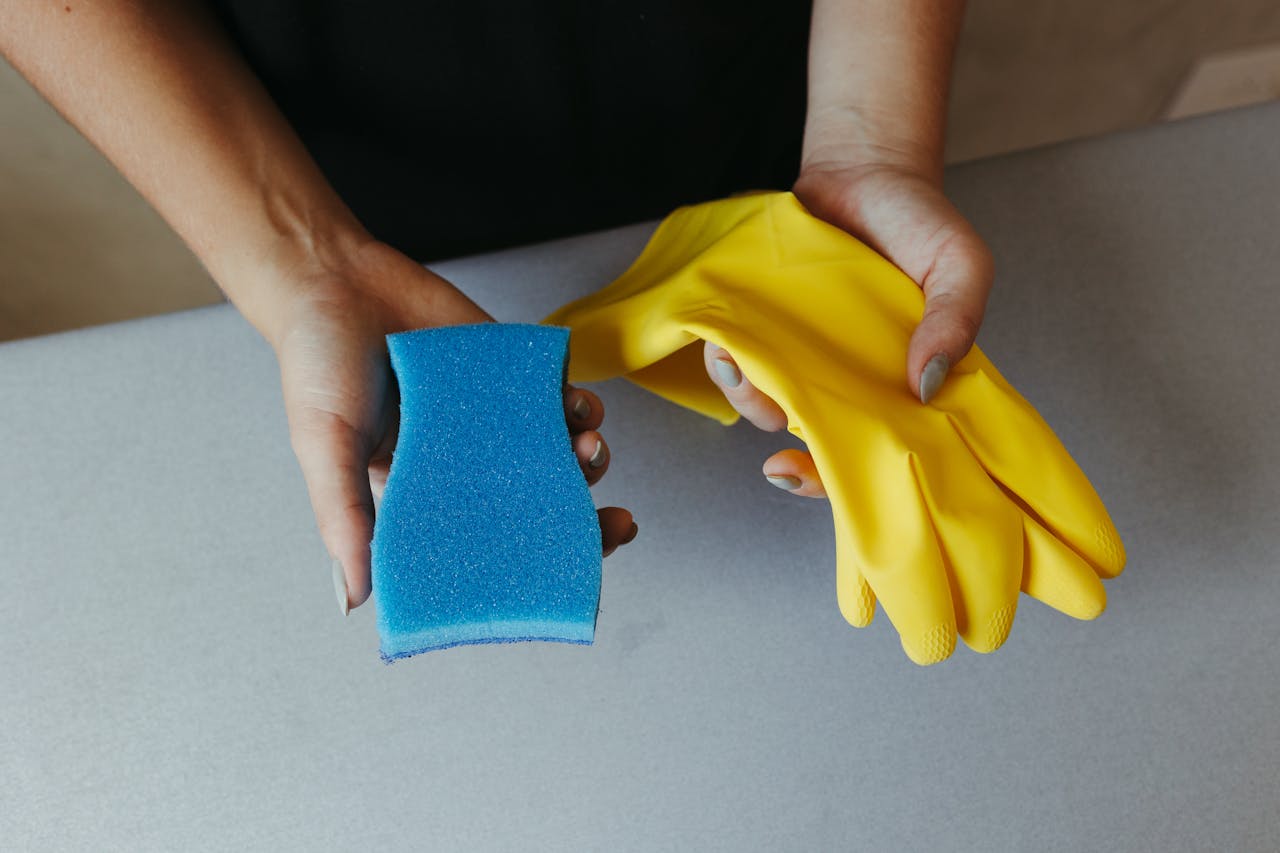 services-01 Close-up of hands holding a blue sponge and yellow rubber gloves on a table.