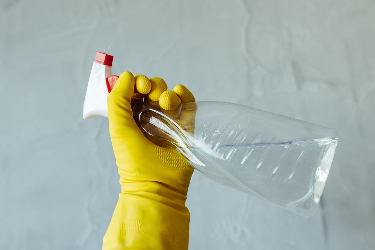 services-02 Close-up of a hand wearing yellow gloves holding a transparent spray bottle, isolated on a plain background.