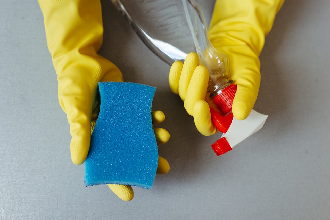 hero-img-01 Hands wearing yellow gloves holding a sponge and a spray bottle.